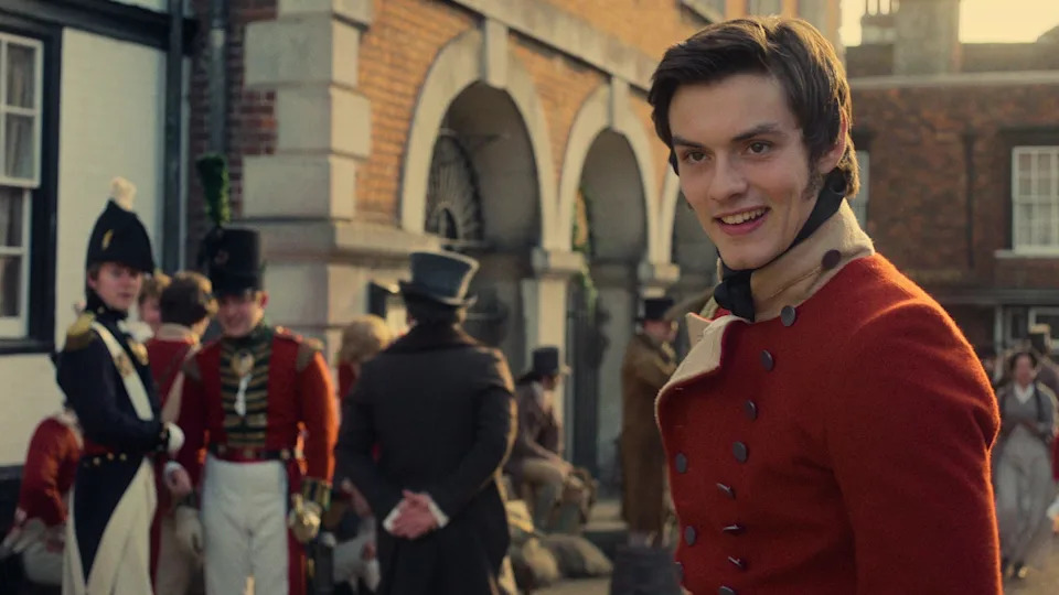 A young man in a red coat smiles, surrounded by historical figures in a lively marketplace setting.