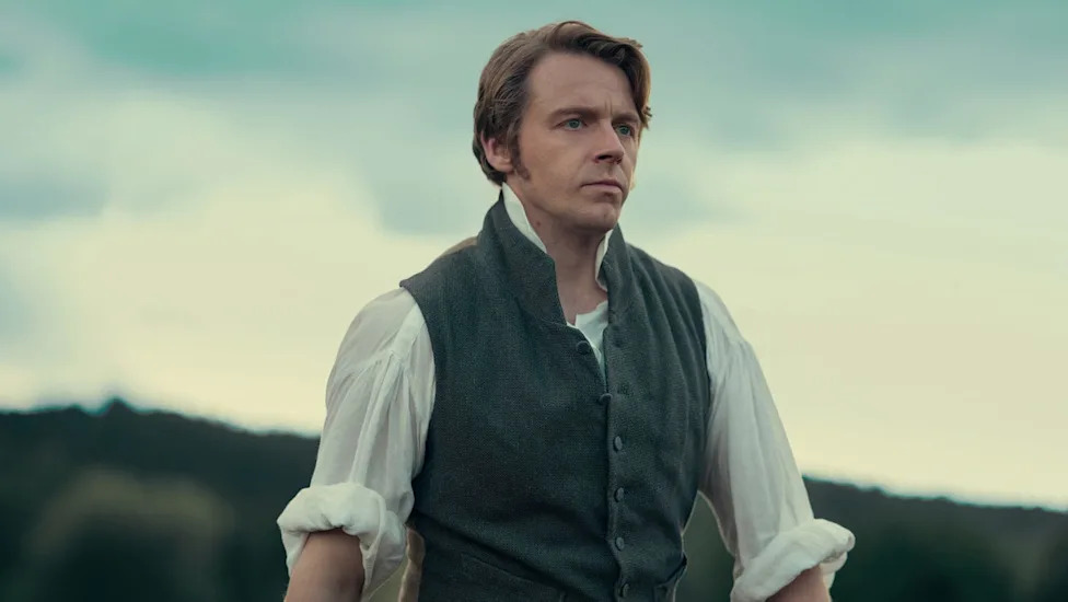 Man in period clothing stands outdoors, looking thoughtfully into the distance against a cloudy sky.