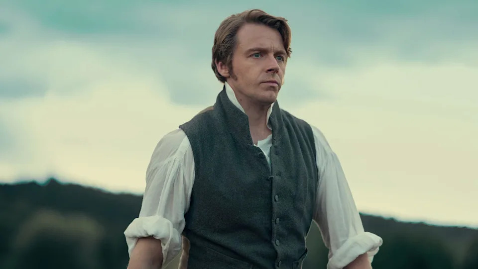 Man in period clothing stands outdoors, looking thoughtfully into the distance against a cloudy sky.