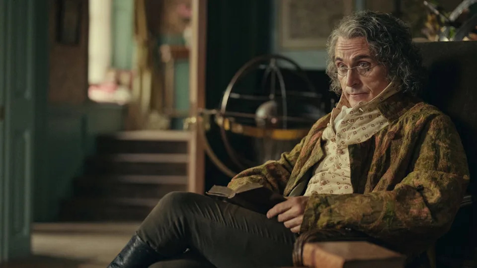 A person with curly hair wearing a patterned coat sits in a chair, holding an open book in a cozy, vintage room.