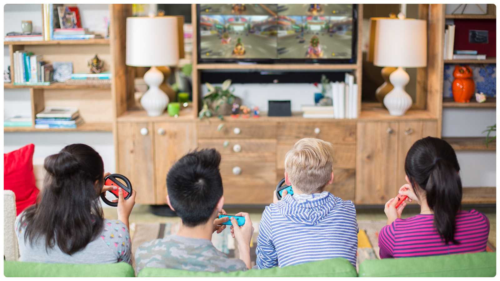 Four people sitting together on a sofa in a living room, holding Joy-Con controllers and playing a Nintendo Switch game on the television.