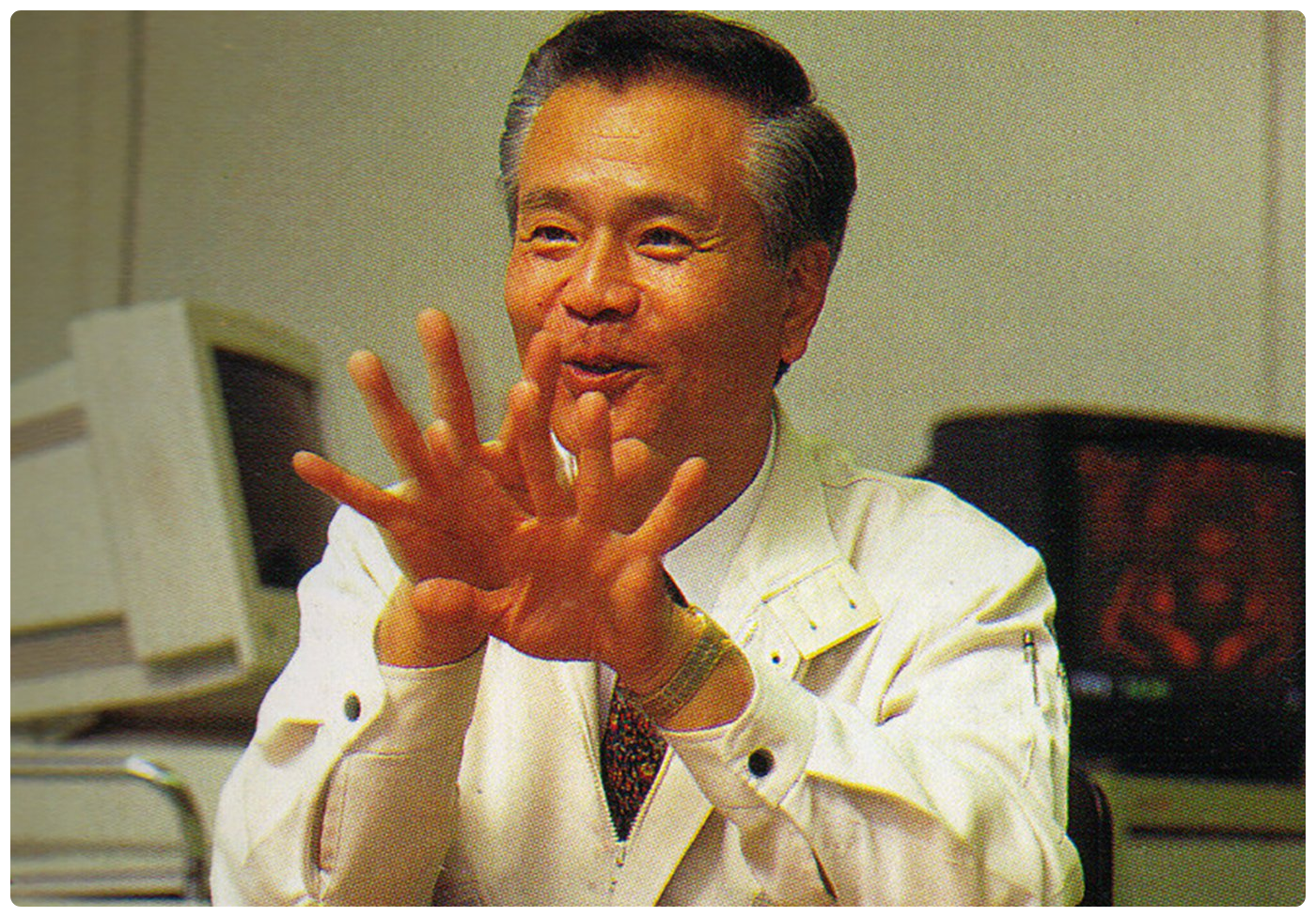 Nintendo designer Gunpei Yokoi smiling and gesturing with both hands raised, seated in front of 1990s desktop computers.
