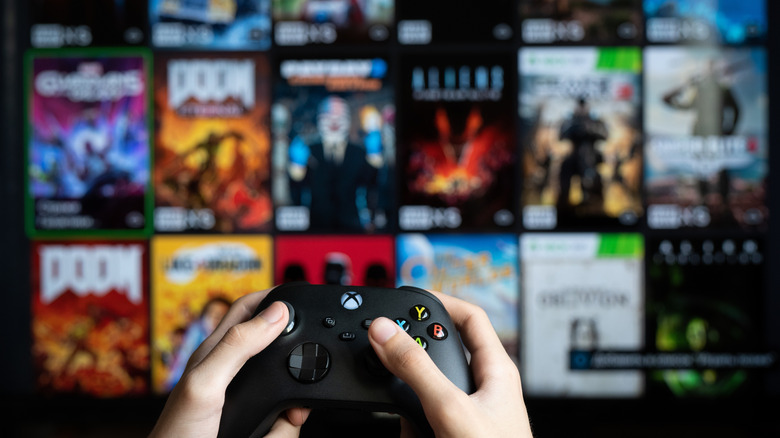 Person holding Xbox controller in front of a TV displaying various Xbox games