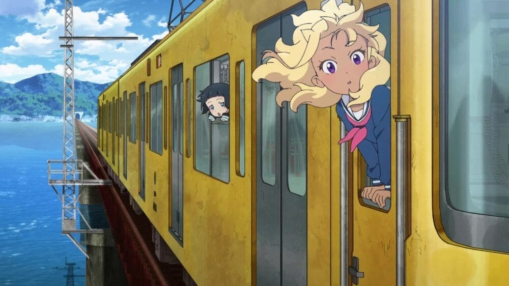 Exterior shot of a yellow train driving down a bridge with blue water underneath. Two teen girls are poking their heads out the windows, hair blowing in the breeze
