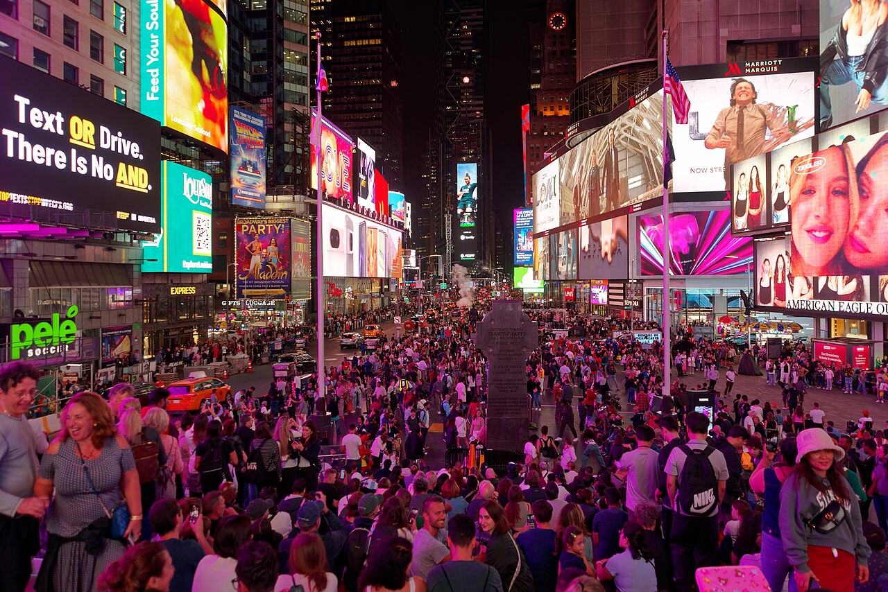 Times Square at night, crowded with people and lit by giant billboards on every side, with the stone Father Duffy monument in the middle of the plaza