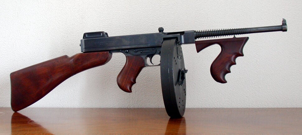 A vintage Thompson submachine gun with a wooden stock, foregrip, and pistol grip rests on a wooden surface. It features a round drum magazine and a simple rear sight against a plain, light-colored wall background.