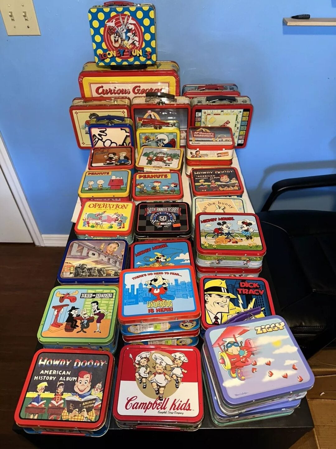 An assortment of colorful vintage lunchboxes stacked on a chair and table against a blue wall. Designs include cartoon characters, superheroes, and iconic brands, showcasing nostalgic themes and varied artwork.