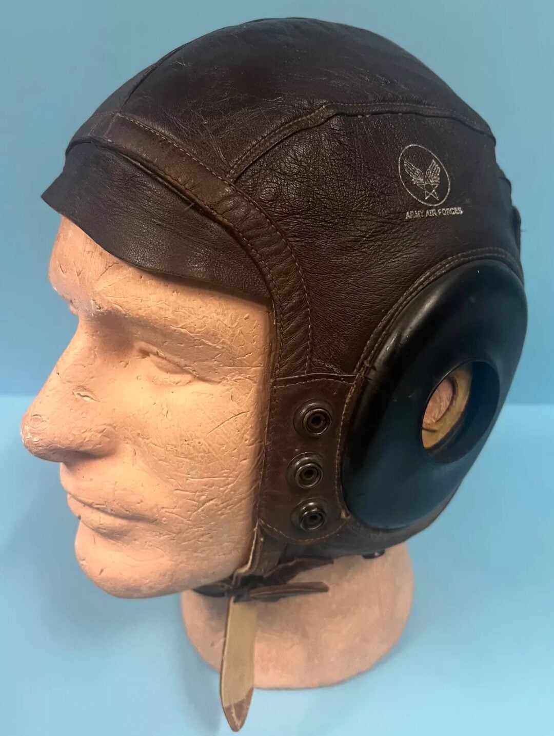 A vintage pilot's cap, made of brown leather, displayed on a mannequin head. The cap features a round black ear covering with multiple rivets and a strap under the chin. A logo and "ARMY AIR FORCES" text are visible on the side.