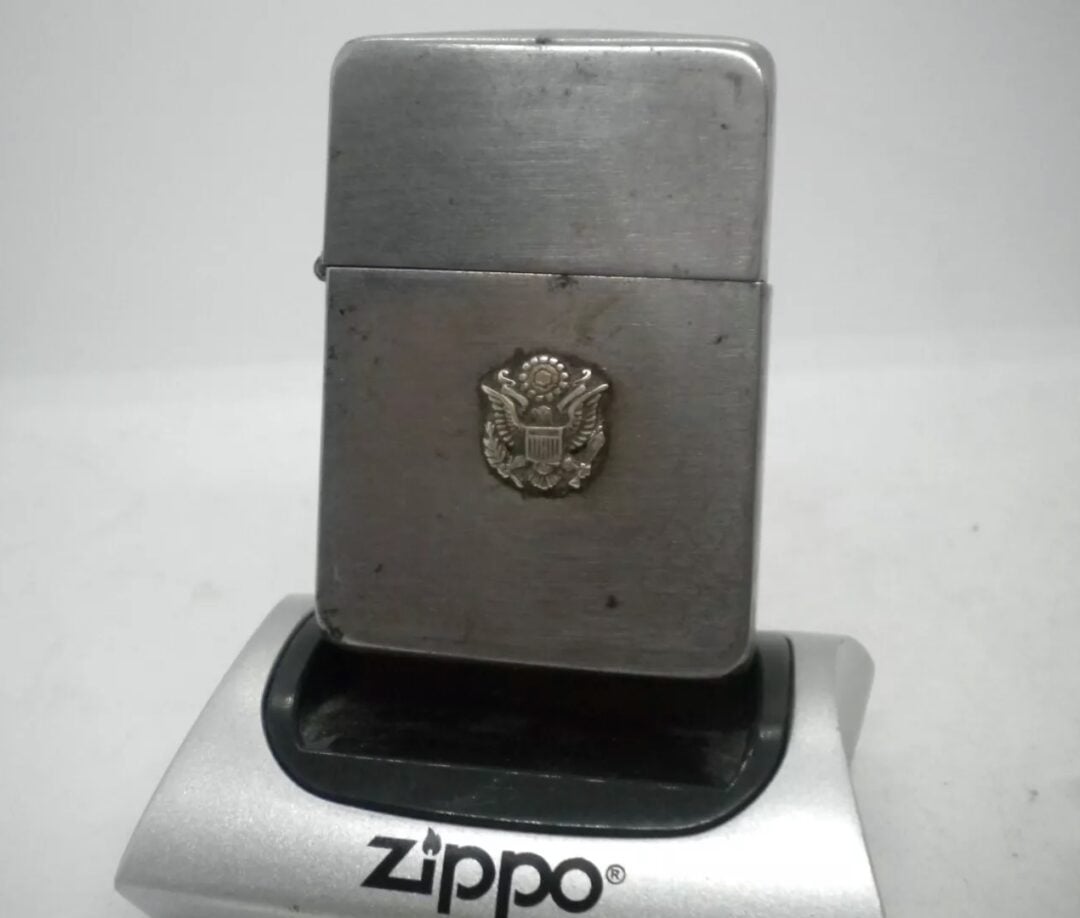 A silver Zippo lighter displayed on a stand with the word "Zippo" on it. The lighter features an embossed emblem resembling a coat of arms, possibly American. The background is plain and white.