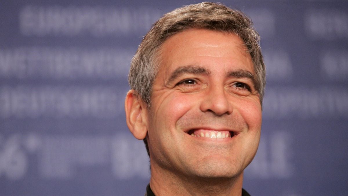 A man with short gray hair and a wide smile stands in front of a blurry blue background with white text.
