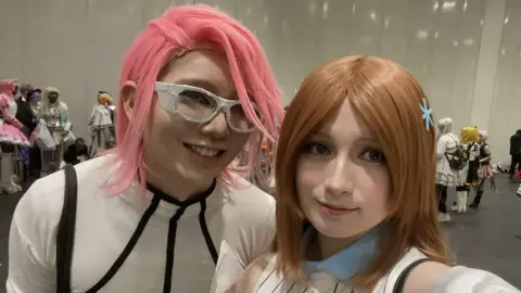 Ruby Ebbs Nicholas on the right wears a white bodysuit with black piping and a pink wig. Ruby wears a blue collar and white striped outfit and a brown wig. They both look at the camera and smile.