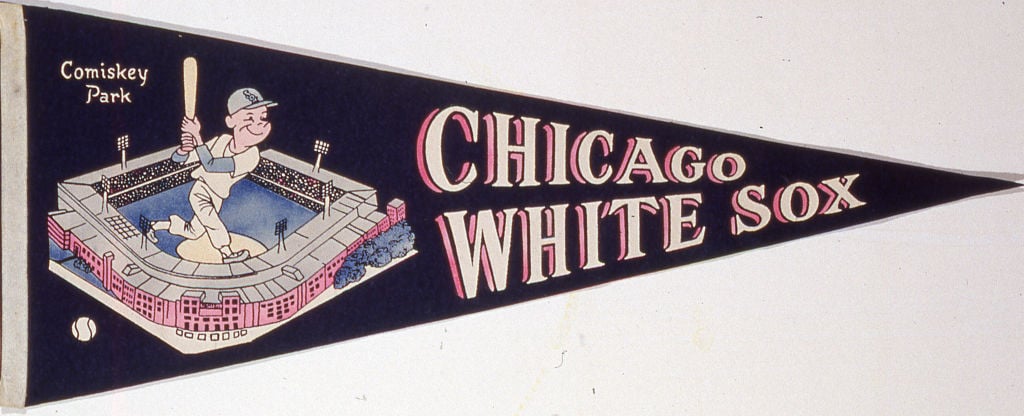 A vintage pennant featuring a cartoon baseball player swinging a bat inside Comiskey Park, with the text "Comiskey Park" above and "Chicago White Sox" in bold letters to the right. The background is dark, highlighting the illustration and text.