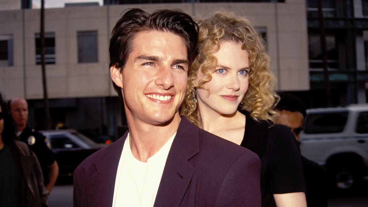 A smiling man in a dark jacket stands in front of a woman with curly blonde hair, outdoors in an urban setting with cars and a building in the background.
