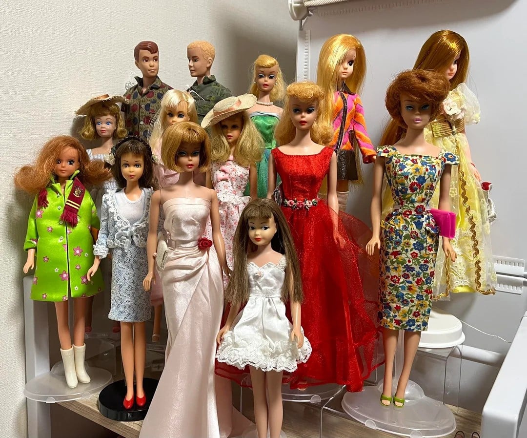 A group of vintage dolls in various styles and colorful outfits, including dresses and hats, are arranged on a shelf. The dolls have different hair colors and styles, showcasing a collection with retro fashion.