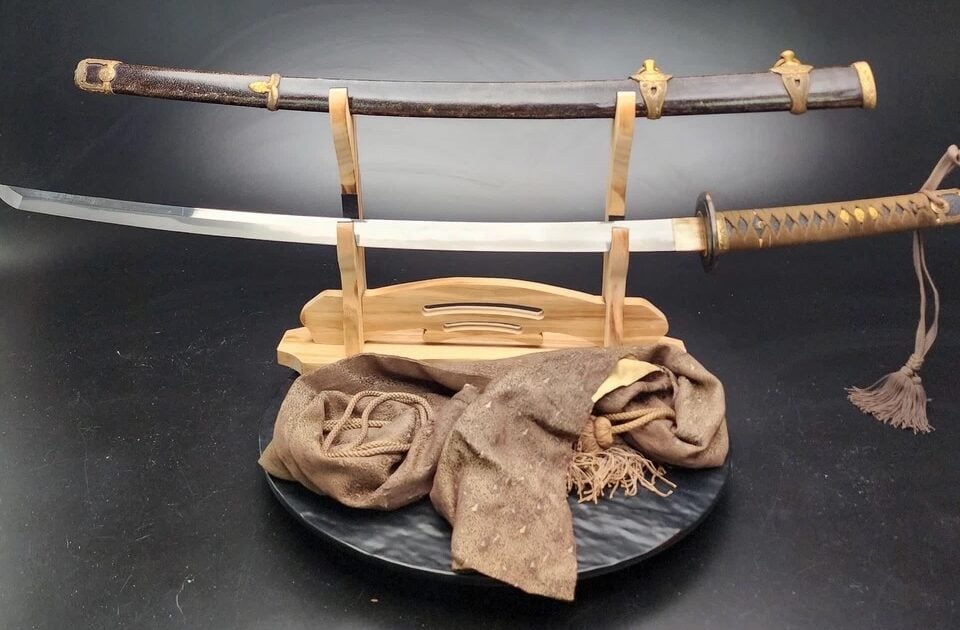 A traditional Japanese katana sword displayed on a wooden stand. The sword has a shiny blade and a decorative handle, with the scabbard displayed above. A brown cloth rests at the base of the stand. The background is dark.