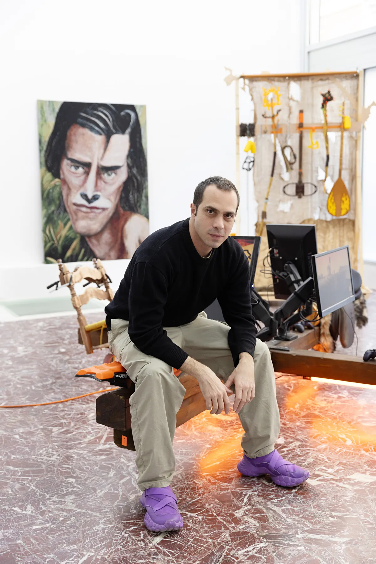 Artist Theo Triantafyllidis sits on a wooden leg of his installation in his studio, an expressionist portrait of a man with black hair  is in the background.