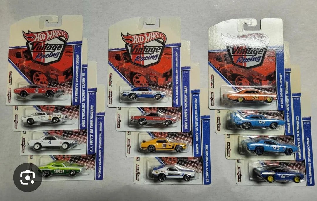 A collection of 13 Hot Wheels Vintage Racing cars in packaging, displayed against a neutral background. The cars vary in color, including white, blue, yellow, green, and orange, with racing details and decals visible on the packaging.