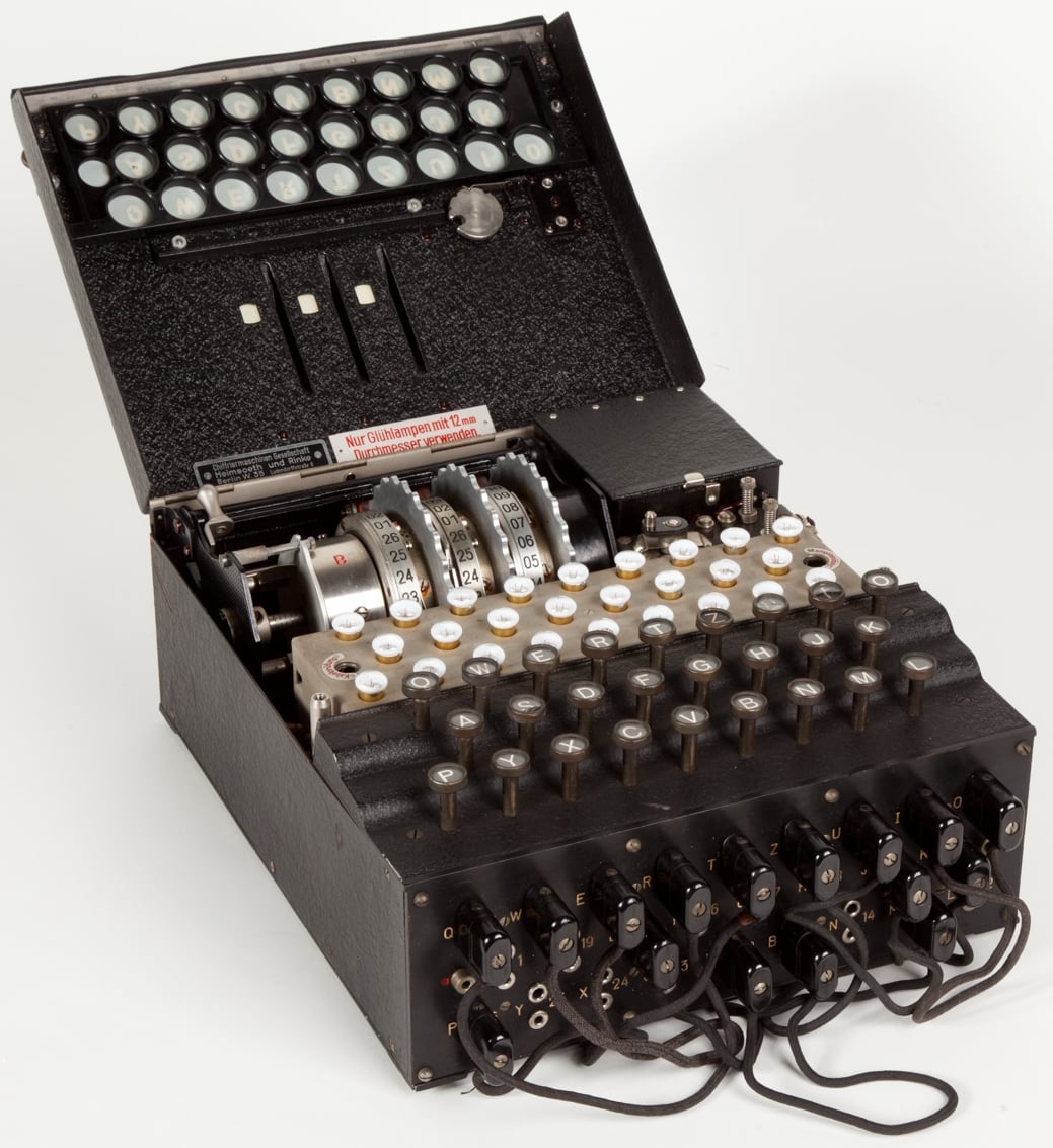 An Enigma machine with its lid open, showcasing a series of rotors, keys, and lamps on a black casing. It has circular keys and a plugboard with cables at the front, used for encryption during World War II.