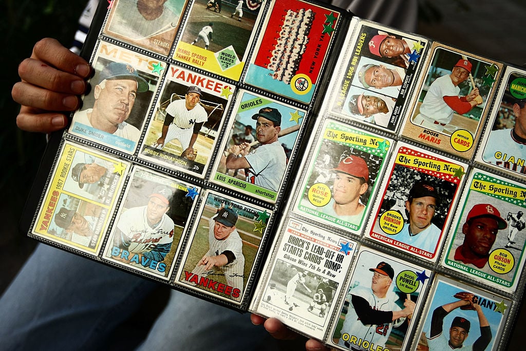 Hands holding a binder filled with vintage baseball cards featuring various players in action and portrait shots. The cards display colorful designs and team names, neatly organized in plastic sleeves.