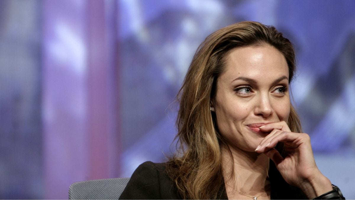 A woman with long brown hair sits, smiling slightly with her hand resting near her mouth. She wears a dark blazer and a delicate necklace. The background is blurred with purple and blue tones.