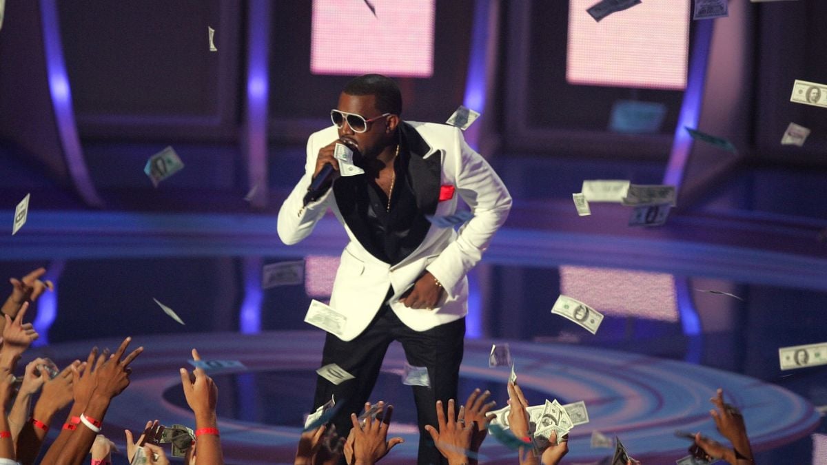 A performer in a white blazer and sunglasses sings on stage, surrounded by falling dollar bills and reaching hands from the audience.