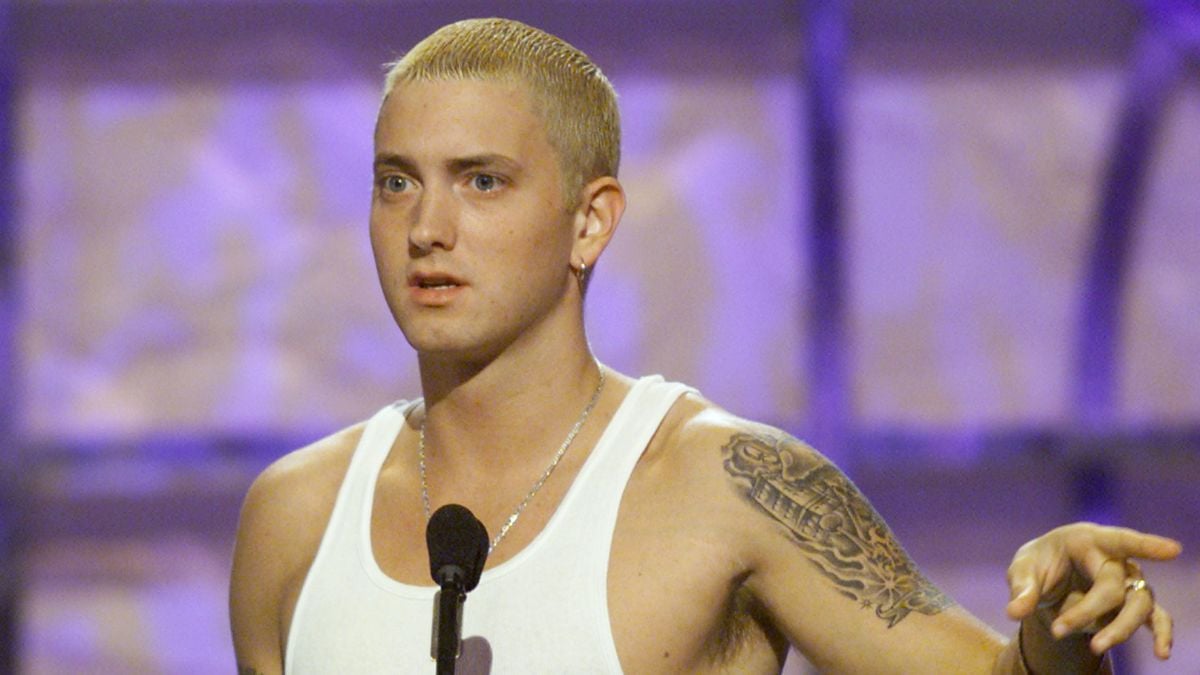 A man with short bleached blond hair and a tattoo on his upper arm speaks into a microphone while wearing a white tank top against a blurred purple background.