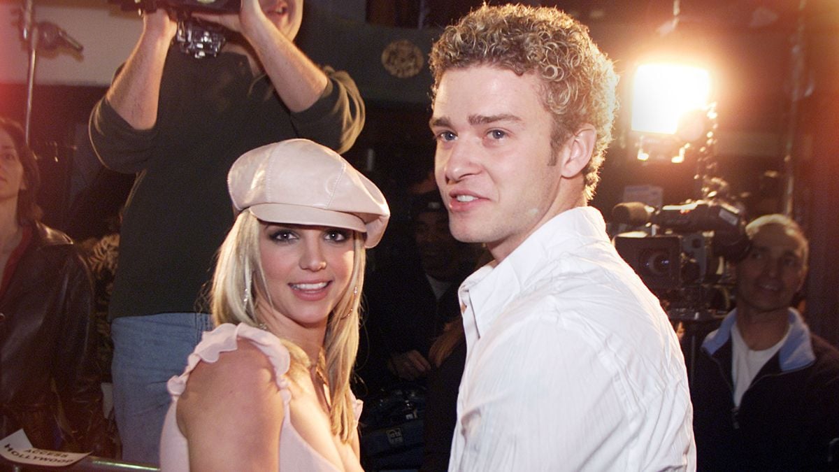 Two young adults pose together on a red carpet at night, surrounded by cameras and lights. The woman wears a pink outfit and hat; the man wears a white shirt. Photographers and camera operators are seen behind them.