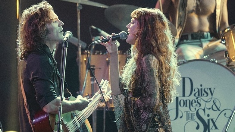 Sam Claflin as Billy strumming guitar on stage next to Riley Keough as Daisy singing into microphone in Daisy Jones and the Six