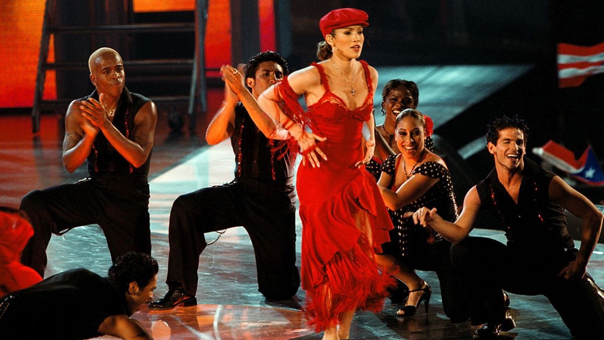 A woman in a red dress and hat dances energetically on stage, surrounded by backup dancers in black outfits, under vibrant lighting.