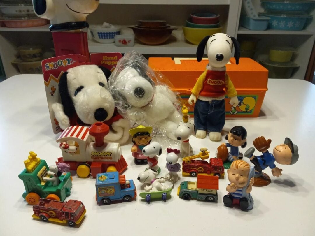 A collection of Snoopy and Peanuts character toys is displayed on a table. It includes stuffed Snoopy dolls, small figurines, and toy vehicles. A bright orange box is in the background, surrounded by shelves with various dishes.