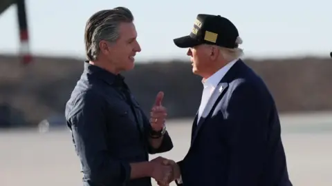 Reuters California Governor Gavin Newsom and US President Donald Trump shake hands on airport tarmac. Trump, on the right, wears a baseball cap.