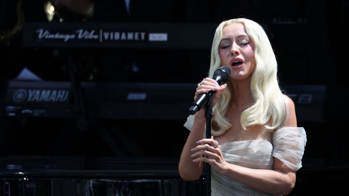 A woman with long blonde hair in a light dress sings into a microphone on stage, eyes closed, in front of a Yamaha keyboard and musicians dressed in black.