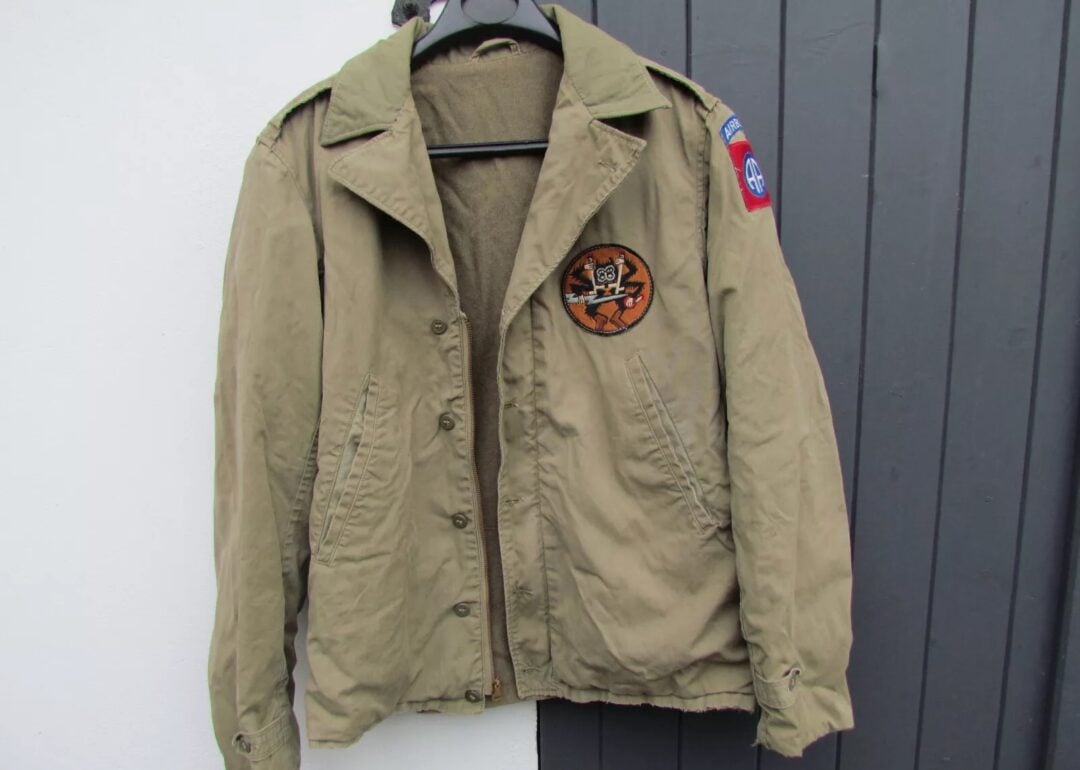 Beige military-style jacket hanging on a black coat hanger. The jacket features a round patch with a tiger emblem on the left chest and a rectangular blue and red patch on the right sleeve. It is displayed against a dark gray background.