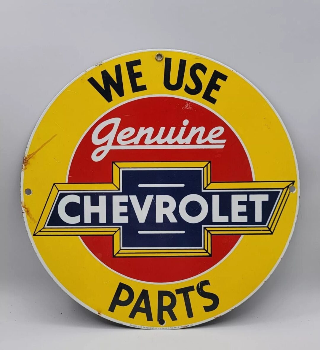 Round vintage sign with a yellow border and red center, featuring the text "WE USE Genuine CHEVROLET PARTS" in bold letters. A classic Chevrolet emblem is prominently displayed in the center.