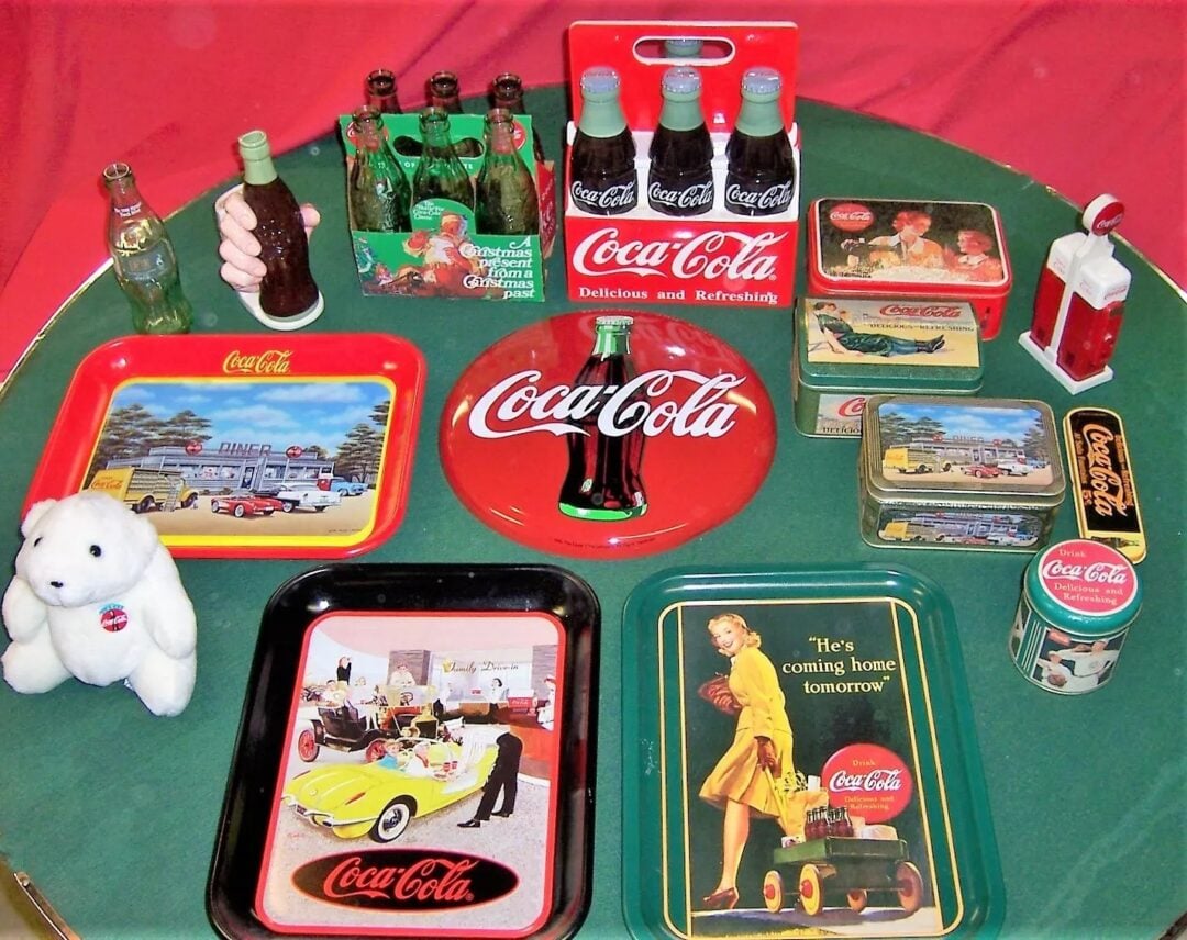 A collection of vintage Coca-Cola memorabilia displayed on a green table. Items include trays, tins, bottles, and a small stuffed polar bear. The background is red, complementing the retro theme with various nostalgic Coca-Cola designs.