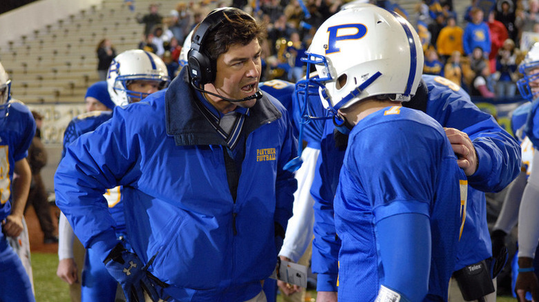 Kyle Chandler as Coach Taylor with blue jacket and and headset talking to player in blue football uniform in Friday Night Lights