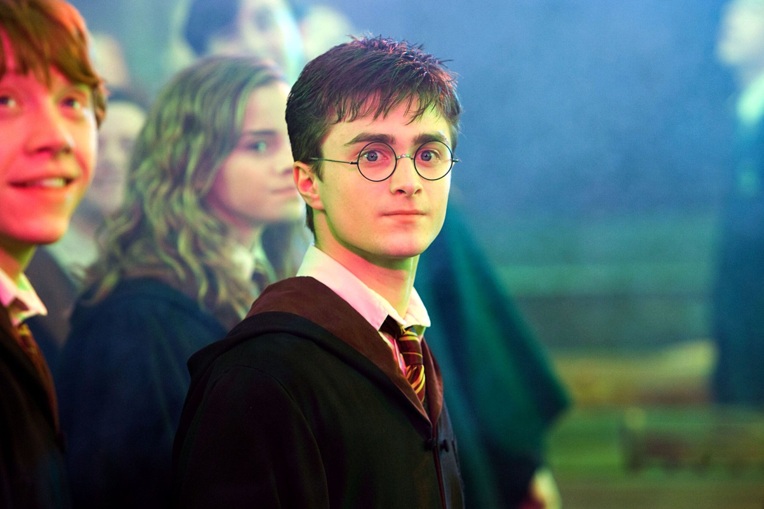 A young man with round glasses and a robe stands in focus, with peers in the background, reminiscent of a school scene