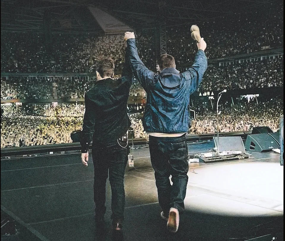 The Gallagher brothers raise their arms on stage to a packed audience.
