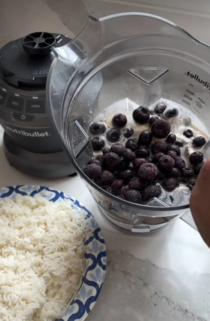 A blender with frozen blueberries and other ingredients next to a paper plate of cooked rice