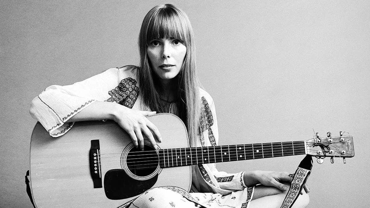 Joni Mitchell sits on the floor with an acoustic guitar in her lap, wearing a white cotton dress.
