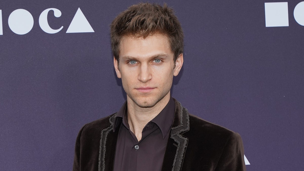 Keegan Michael wearing a dark jacket and button-down shirt poses in front of a purple event backdrop.