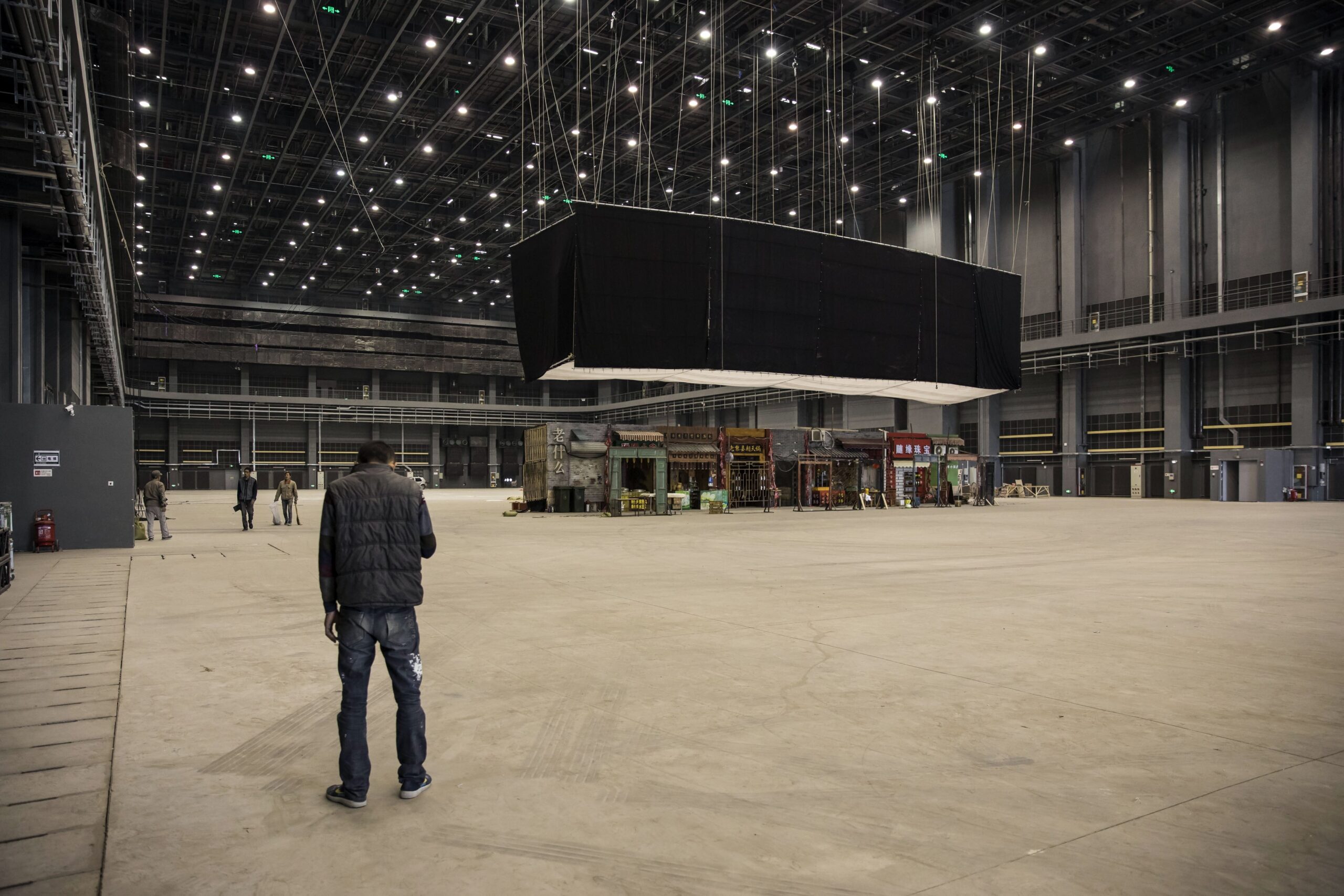 Large empty set with solitary person. High ceiling, suspended screen above, few structures in the background. Film production setting visible